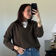 A woman takes a mirror selfie in a home setting, wearing a brown knit sweater, blue jeans, and brown slip-on shoes. A white turtleneck is visible underneath the sweater.